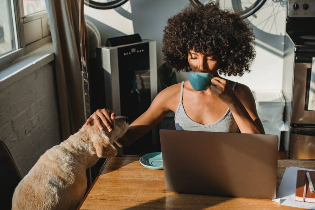 woman at the kitchen table using her laptop, drinking coffee, and petting her dog