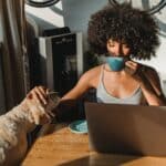 woman at the kitchen table using her laptop, drinking coffee, and petting her dog